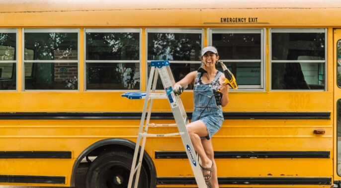 learner in front of a school bus on a ladder holding a drill