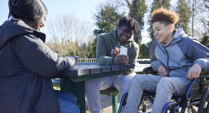 Teenage Girl In Wheelchair With Friends Looking At Social Media On Mobile Phones In Park