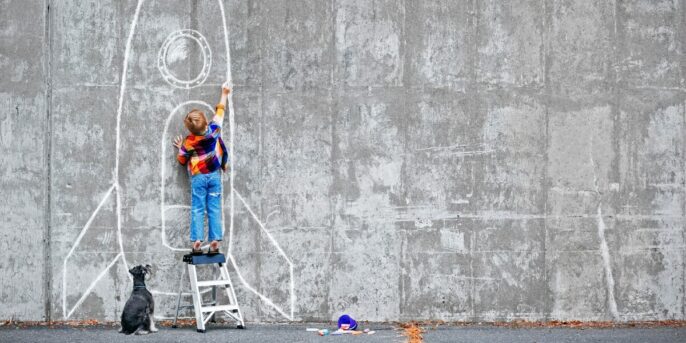 young learner drawing a rocket ship with chalk on a wall with a dog