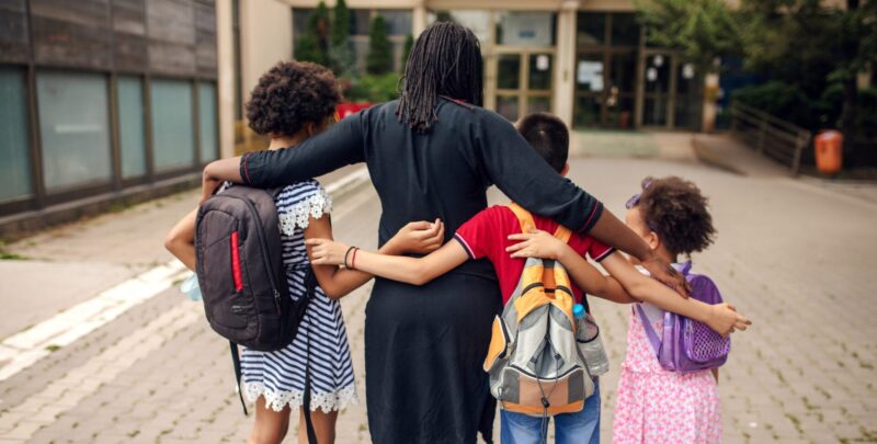 Photo of a mother with her arms around three children, sending them off to school and into the world.